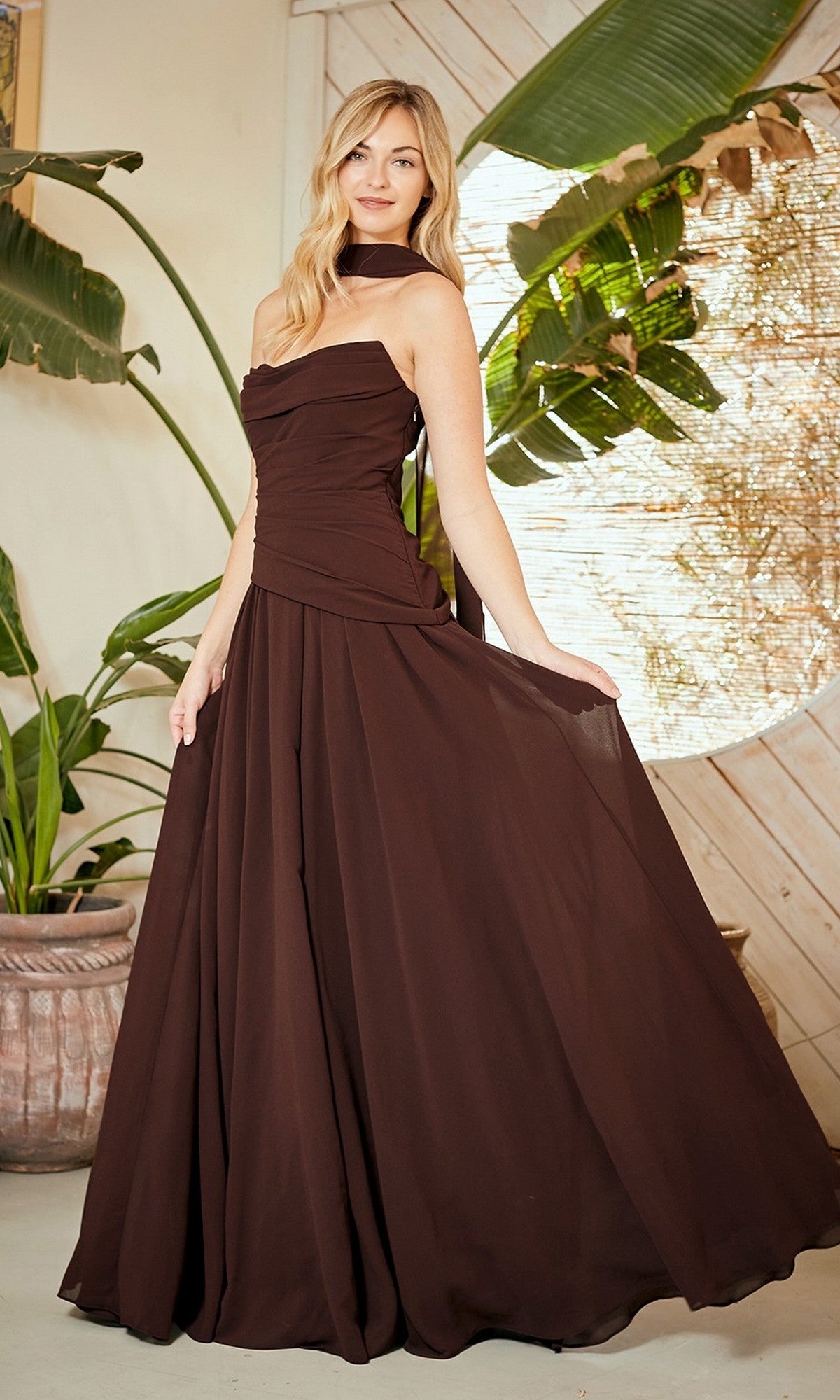 A woman in a brown Amelia Couture 6133 long prom dress with a strapless chiffon design and corset back poses indoors by green plants and a light wood wall.